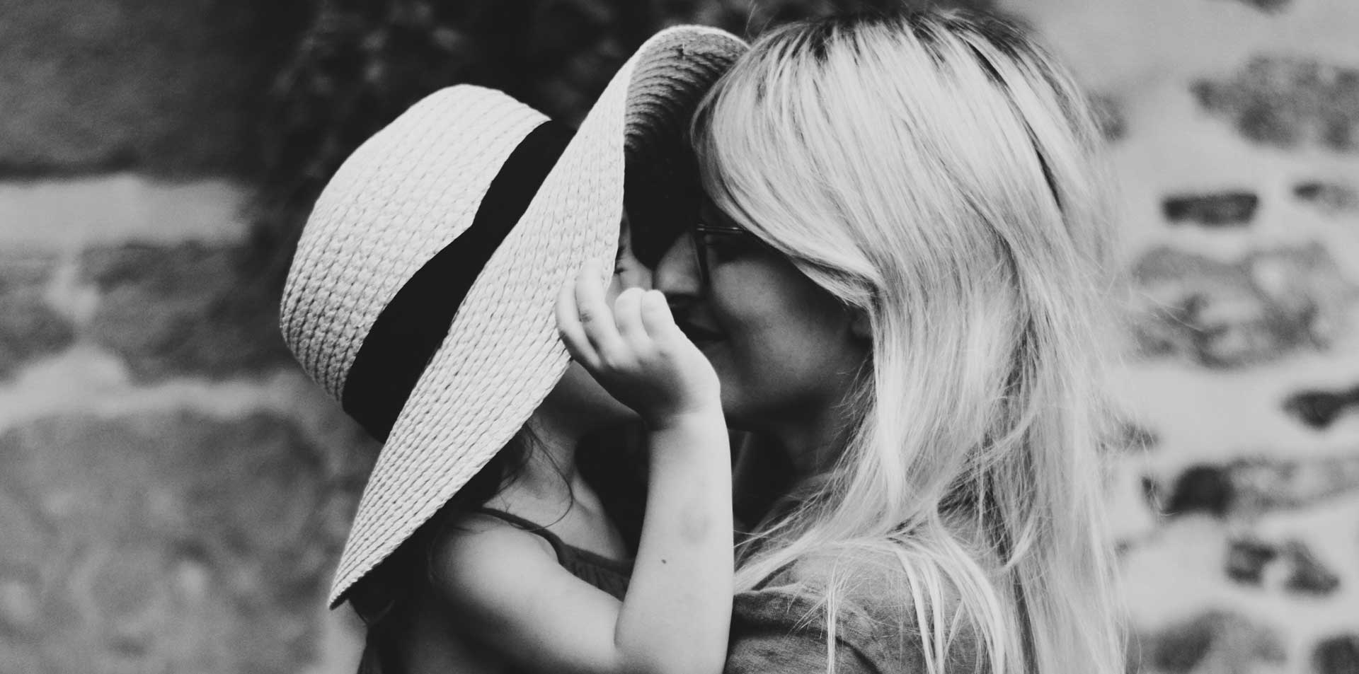 black and white photo of mother with long hair holding child wearing large hat close to her face in front of brick wall